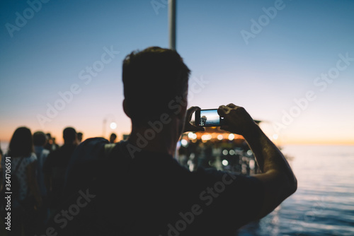 Back view of man taking picture of evening scenery near sea using modern mobile phone camera, male tourist making photo in night twilight via contemporary application for photographing in dark.