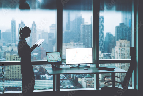 Silhouette of female person hold in hands touch pad while standing near office desktop with Mock up PC computer screen front panoramic city view in window.Trade manager workspace with financial graphs photo