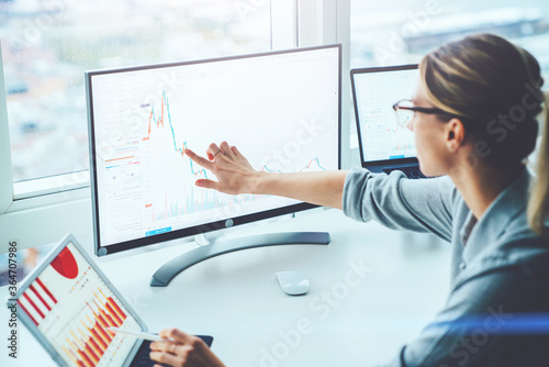 Fotografi Back view of business woman sitting at desktop front PC computer with financial graphs and statistics on monitor