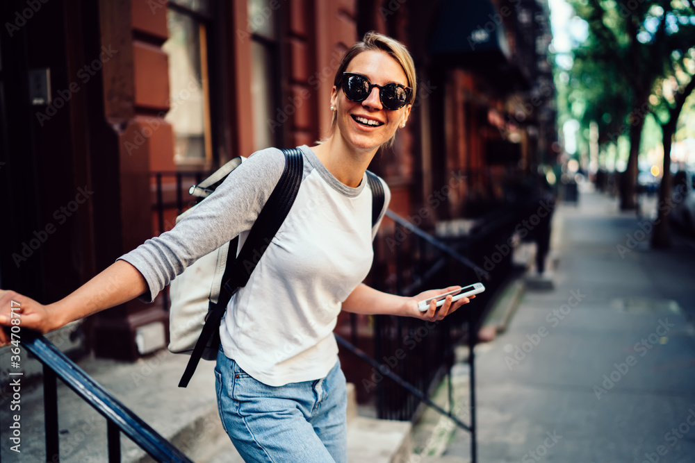 Half length portrait of cheerful young woman leaving building laughing holding telephone,smiling hipster girl in sunglasses spending weekends actively in  strolling in downtown with smartphone.