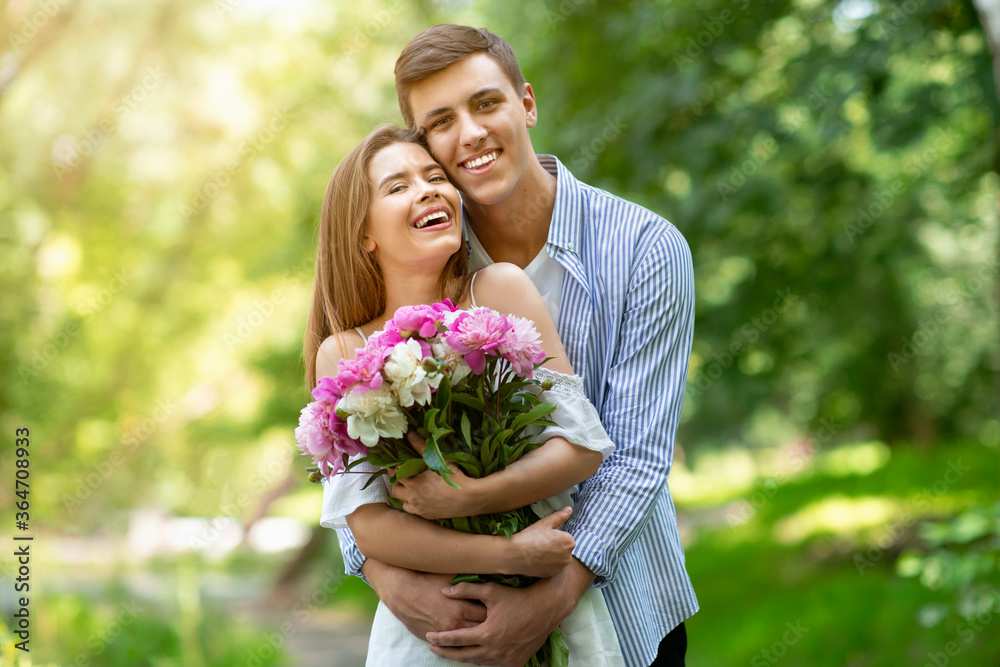 First anniversary gift. Millennial married couple with bouquet of flowers hugging at park