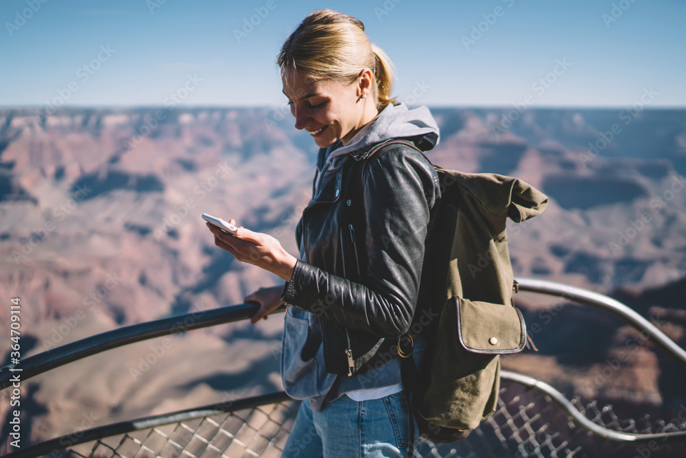 Fototapeta premium Smiling female explorer reading message on telephone satisfied with mobile connection during hiking tour,cheerful girl wanderlust using cellular chatting standing on viewpoint in Grand Canyon.