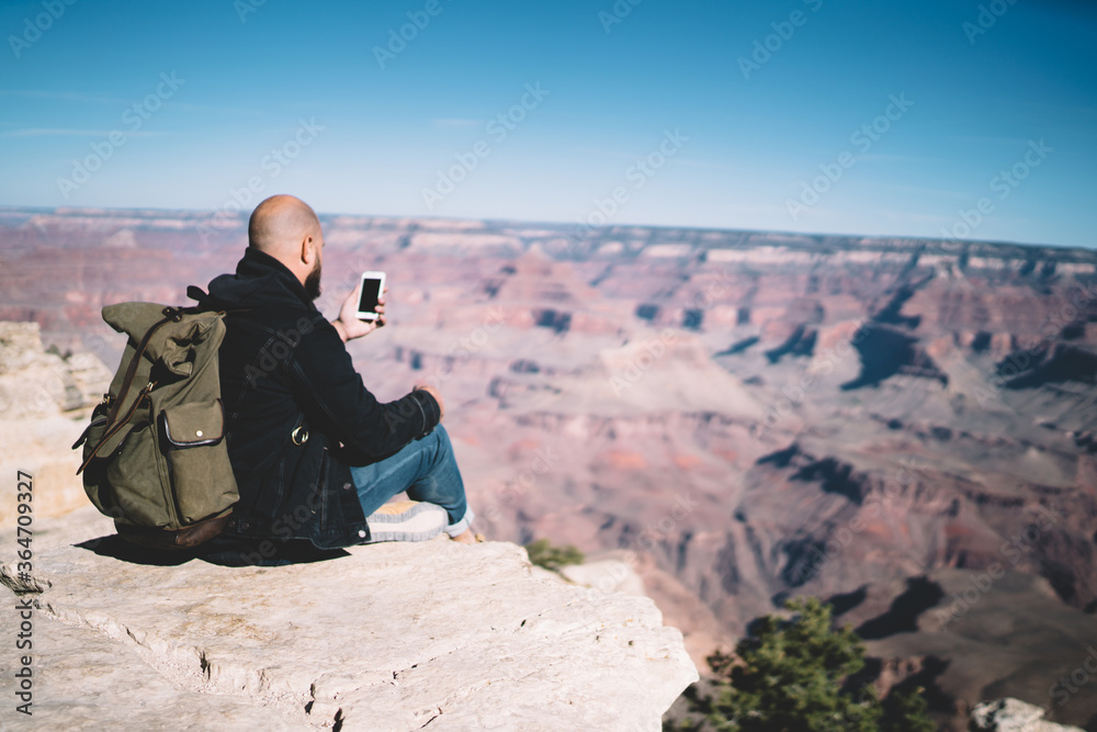 Back view of male explorer wanderlust using smartphone for shooting video for travel blog sitting on high rock in mountains during trip, millennial hipster guy tourist making selfie during hiking tour