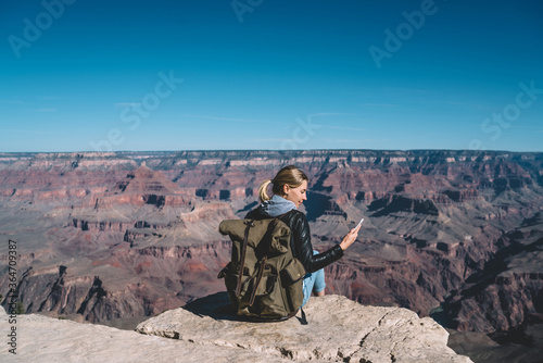 Female traveler holding smartphone checking mobile data connection sitting on mountain top in Grand Canyon National Park, hipster girl enjoying hiking with good network coverage for sending messages