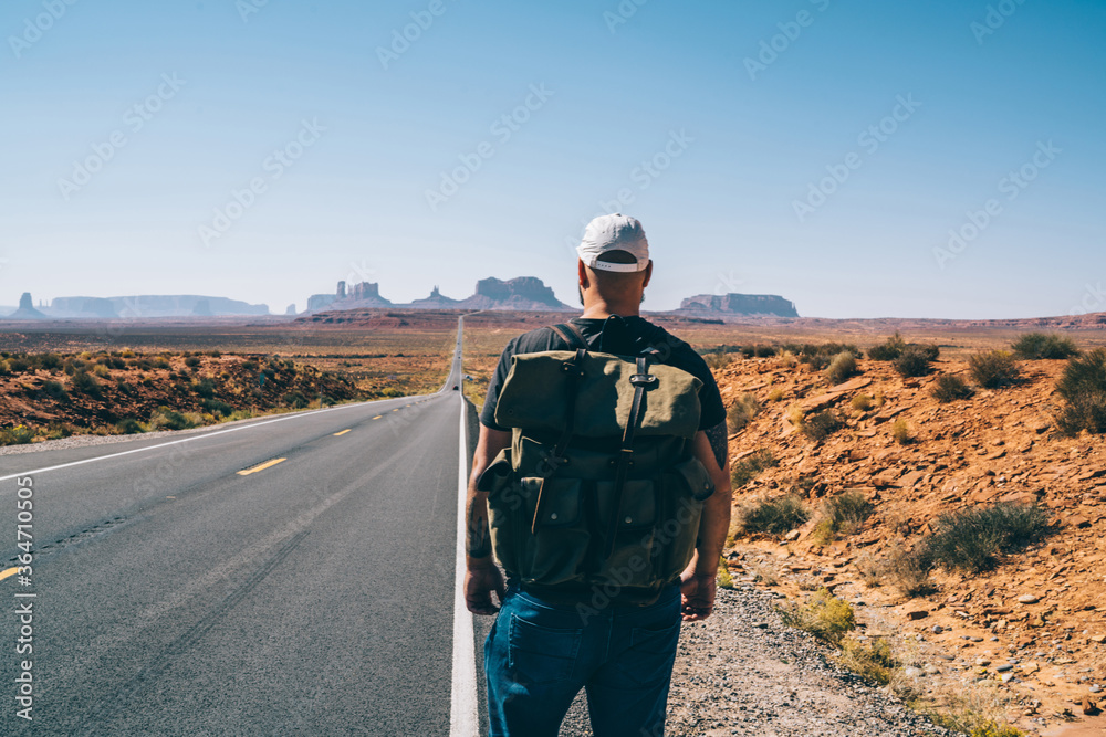 Young hipster guy with rucksack walking near asphalt road lead to National park on southwest, back view of male traveler hitchhiking exploring America having journey to famous landmarks at summer time Young hipster guy with rucksack walking near asphalt road lead to National park on southwest, back view of male traveler hitchhiking exploring America having journey to famous landmarks at summer time