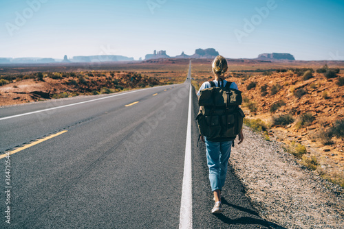 Back view of hipster girl wanderlust walking on asphalt road in wild lands on southwest getting to National park by foot, female tourist with rucksack having journey in Arizona hitchhiking on the way