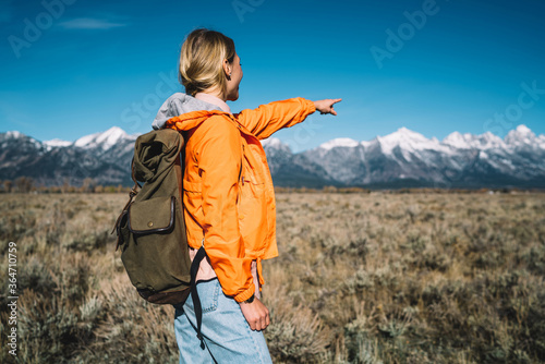 Young female tourist showing beautiful landscape in front of her enjoying discover wild nature, hipster girl with backpack pointing on snow-capped mountains getting there during hiking wanderlust trip