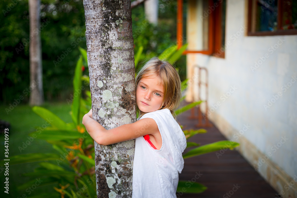 Portrait. Charming little girl standing and holding the tree, wearing white dress. Cute face. Spending time outdoor in tropical garden. Childhood concept. Portrait. Charming little girl standing and holding the tree, wearing white dress. Cute face. Spending time outdoor in tropical garden. Childhood concept.