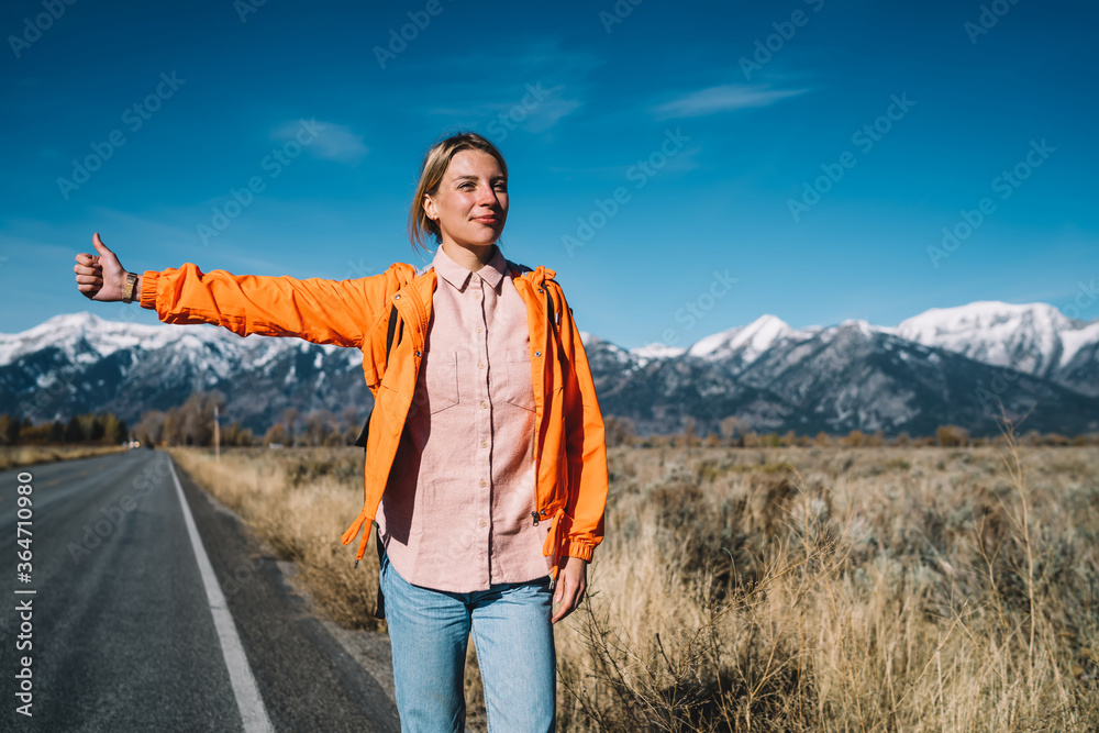 Positive hipster girl waiting for car on asphalt road in national park traveling via auto stop,smiling female wanderlust standing on highway raising hand sign for hitchhiking discover nature
