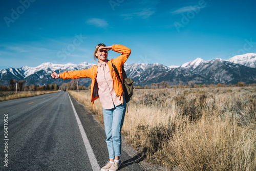 Smiling female hitchhiker raising hand on highway waiting for car getting to destination in mountains,excited cheerful girl wanderlust traveling on autostop explore wild nature on active weekends.