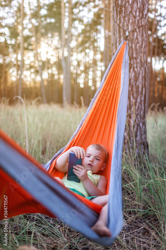 Cute boy having rest and playing on smartphone in hammock in the forest during local vacation