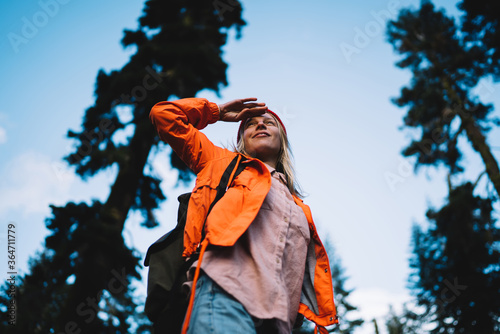 Smiling young female traveler in orange jacket observing route for hiking trails in forest environment, cheerful girl wanderlust enjoying vacation on exploring wild nature in wood with rucksack.