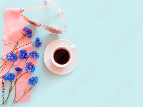 Top view of a cup of coffee with blue cornflowers, pink sunglasses and pink silk ribbon on a pastel blue background. Summer beach flat lay. Lifestyle, copy space, mock-up.