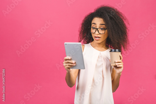 Young american student girl with curly african hair holding digital tablet and coffee or tea over isolated pink background with copy space for text, logo or advertising.