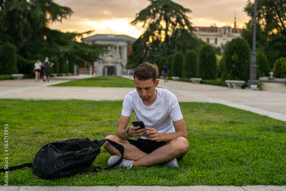 Fototapeta premium Photo of a young and attractive man with a white t-shirt sitting on the grass checking his phone