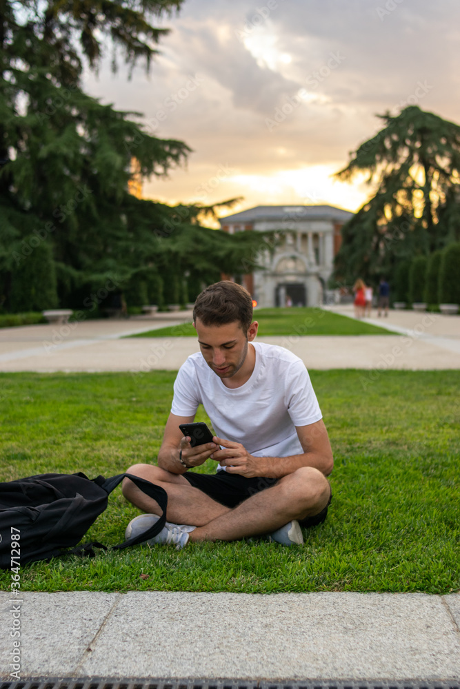 Fototapeta premium Photo of a young and attractive man with a white t-shirt sitting on the grass checking his phone