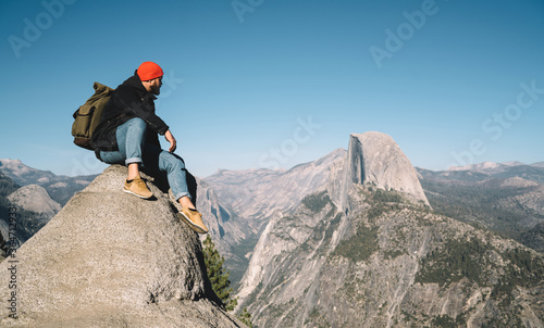 Young hipster guy enjoying wanderlust hiking tour getting to mountain top for observing breathtaking view,male traveler sitting on rocky cliff over abyss enjoying exploring wild environment in usa