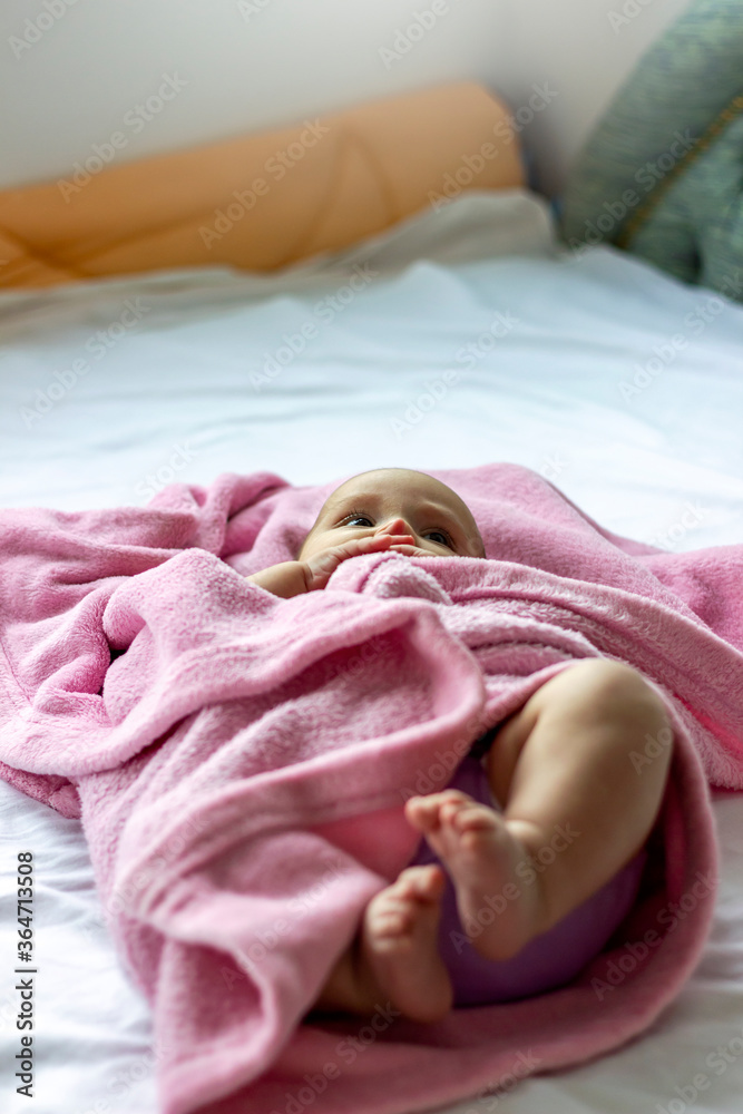 Image of smiling baby girl lying on a bed wrapped in a pink blanket looking at her mother.Adorable four months old caucasian baby girl lying on bed on back with eyes open biting fingers,looking away.