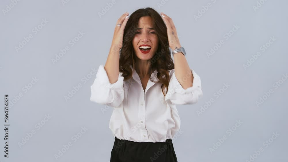 Brunette model in white shirt and black skirt is in despair, screaming and grabbing her head while posing against gray background. Close up Brunette model in white shirt and black skirt is in despair, screaming and grabbing her head while posing against gray background. Close up
