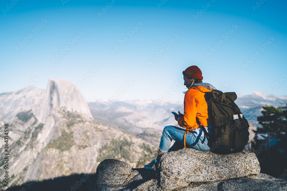 Obraz premium Female tourist with backpack sitting on mountain peak sending text messages on smartphone via good data connection,hipster girl enjoying beautiful landscape of high rock cliffs reading mail on mobile