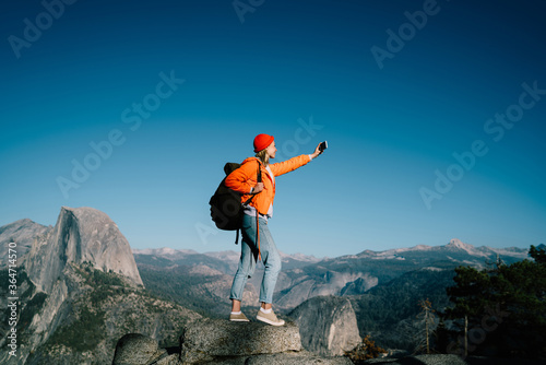 Young female travel blogger taking selfie on smartphone camera during hiking tour in mountain,girl wanderlust with backpack posing for photo on telephone standing on breathtaking scenery 