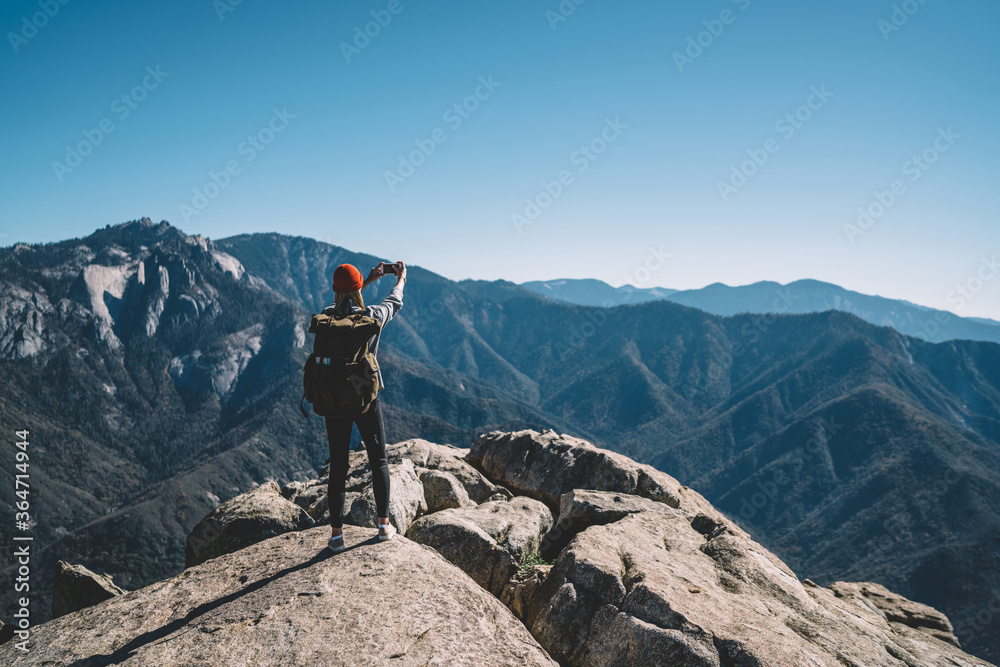 Back view of hipster girl wanderlust using smartphone camera for making selfie on mountain peak, female travel blogger taking picture on telephone on breathtaking landscape scenery of national park.