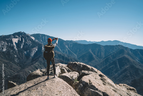 Back view of hipster girl wanderlust using smartphone camera for making selfie on mountain peak, female travel blogger taking picture on telephone on breathtaking landscape scenery of national park.