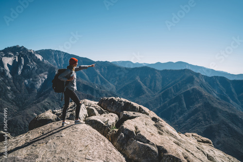 Young female traveler with backpack pointing on breathtaking scenery of mountains standing on hill top, girl wanderlust observing landscape view from rock shoring distance to destination in tour.