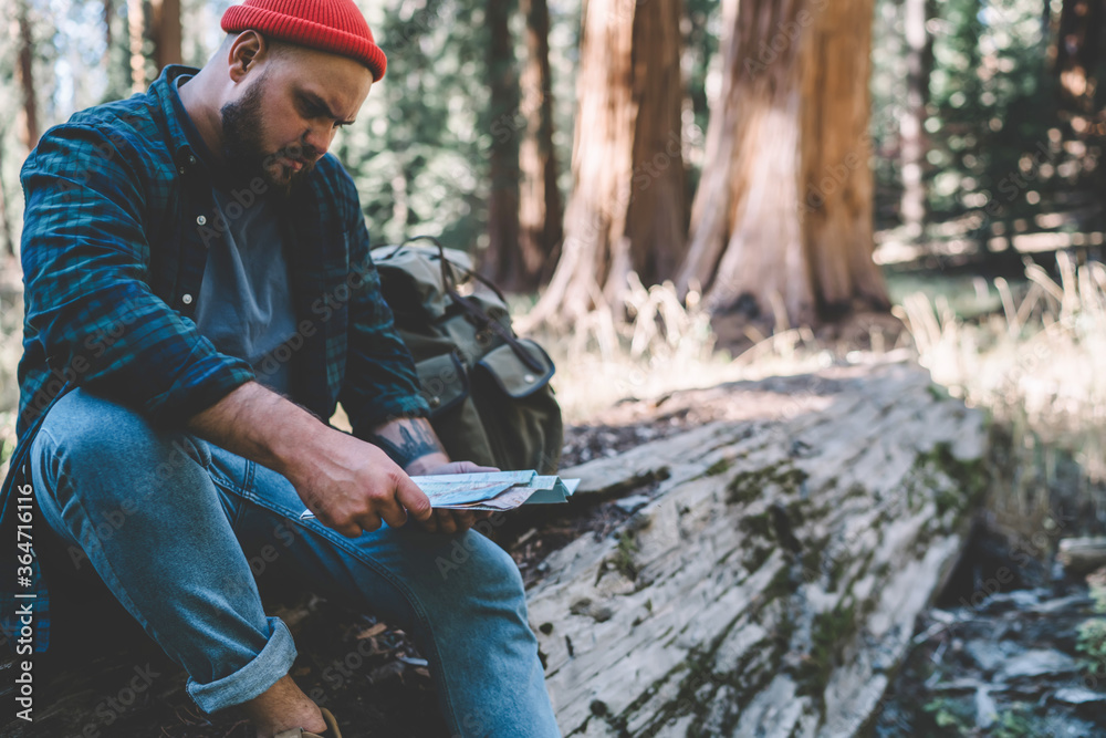 Pensive hipster guy pondering on choosing best route for getting to destination making rest during hiking, male traveler wanderlust sitting in forest reading map for navigate explore wild environment