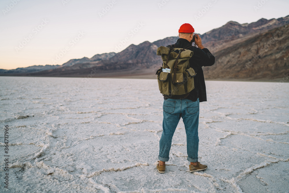 Back view of male traveler with backpack making picture of landscape in desolate lands of desert, hipster guy with rucksack having expedition to Badwater basin taking photo of breathtaking scenery.