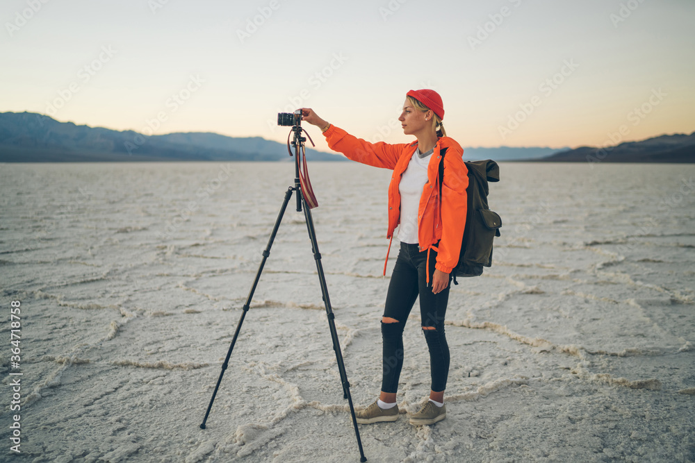 Girl in touristic orange jaket standing with tripod for shooting video of wild environment of death valley, skilled female photographer taking photos of landscape and desolate surface on moder camera