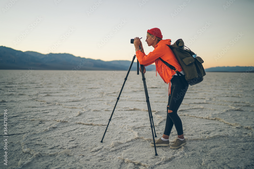 Female photographer standing with tripod in desert dry lands shooting sunset and landscape on modern equipment, skilled hipster girl making photos on camera at twilight in badwater basin national park