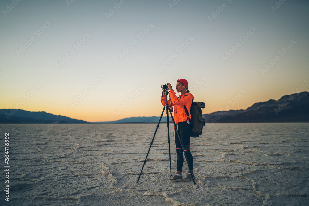 Skilled female photographer with equipment taking photos on camera and tripod during evening in desert environment, young woman wanderlust shooting video of sunset  in expedition in Badwater .