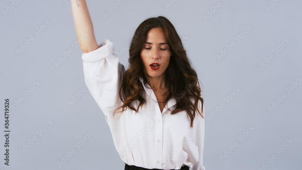 Brunette female in white shirt is looking happy, smiling and dancing funny dances while posing against gray studio background. Close up Brunette female in white shirt is looking happy, smiling and dancing funny dances while posing against gray studio background. Close up