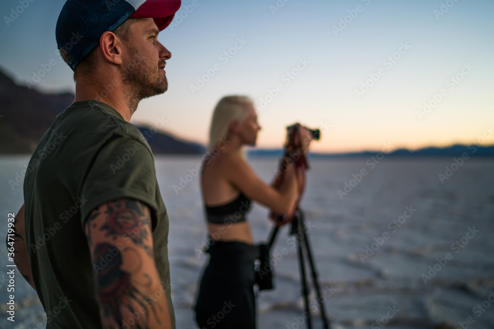 Obraz premium Cropped image of handsome male traveler on frontage standing near woman shooting video of sunset in desert,male and female friends taking photos during journey in Badwater basin national park.