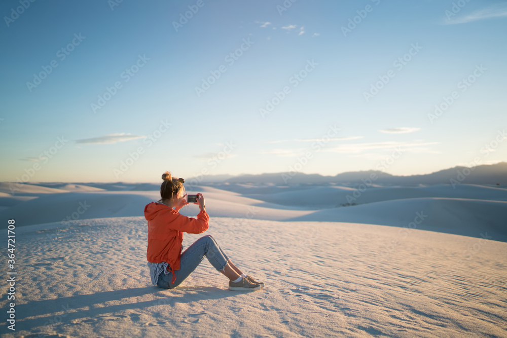 Young hipster girl using smartphone camera for taking picture of white dunes desert at susnet, woman photographing on telephone camera exploring nature view  during journey to famous landmark of USA