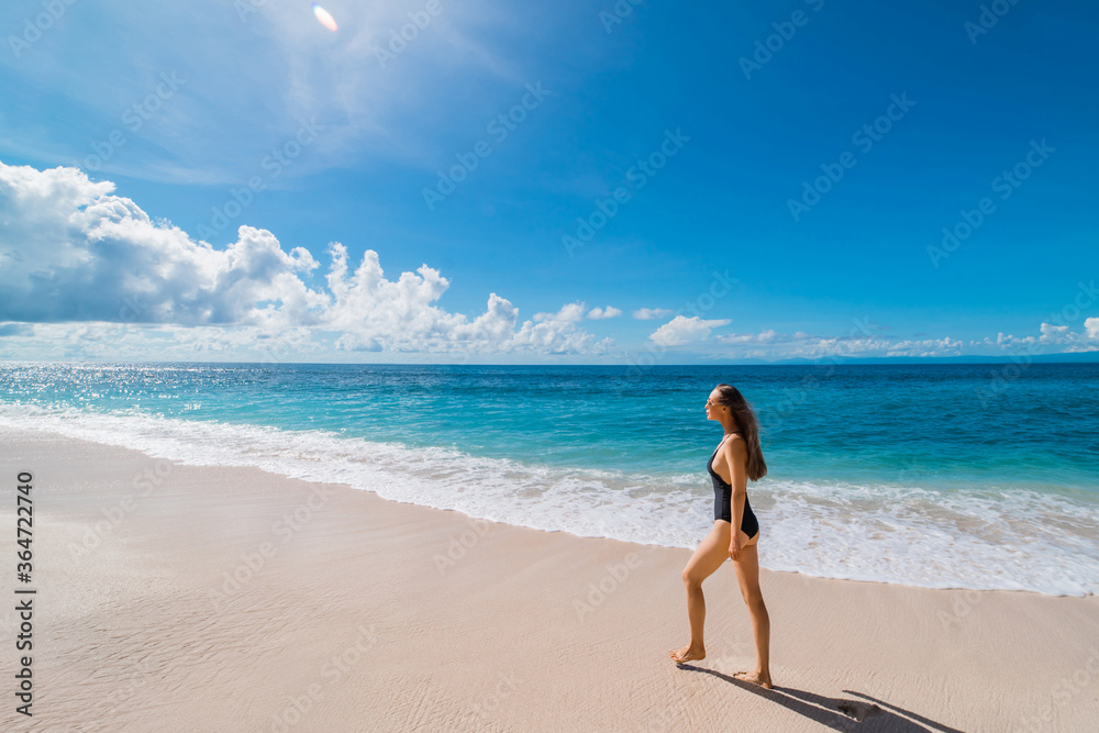 A young tourist girl walks on an empty beach on the island of Bali. A sexy girl in a black swimsuit is photographed with a wide-angle lens walking barefoot