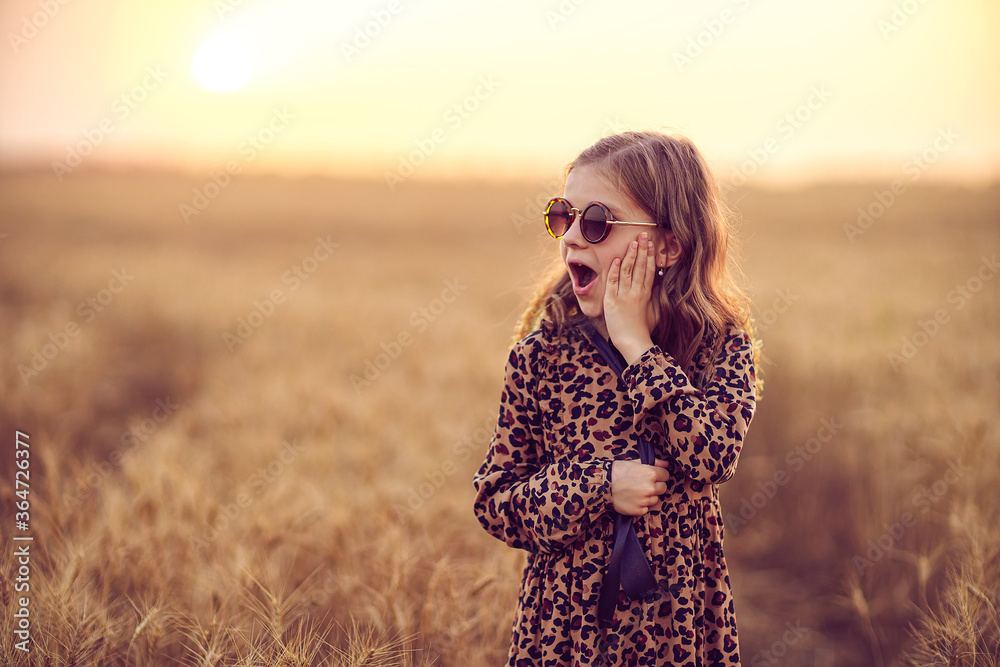 Fashion photo of a little girl in leopard print dress, sunglasses and straw hat at the evening wheat field.