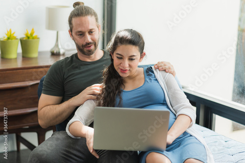 Beard man and his pregnant wife watching movie on laptop feeling happy while sitting on sofa in the home. Family concept.