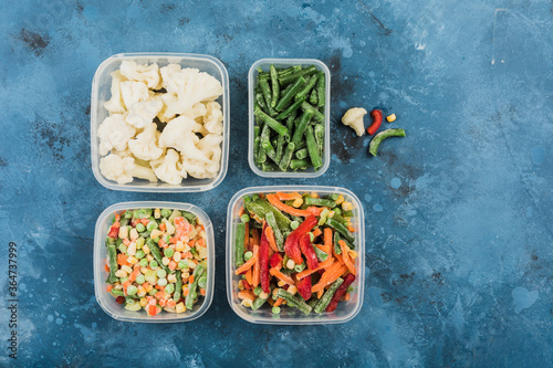 Frozen vegetables: a mix of vegetables, string beans and cauliflower in different plastic containers for freezing on a blue background.