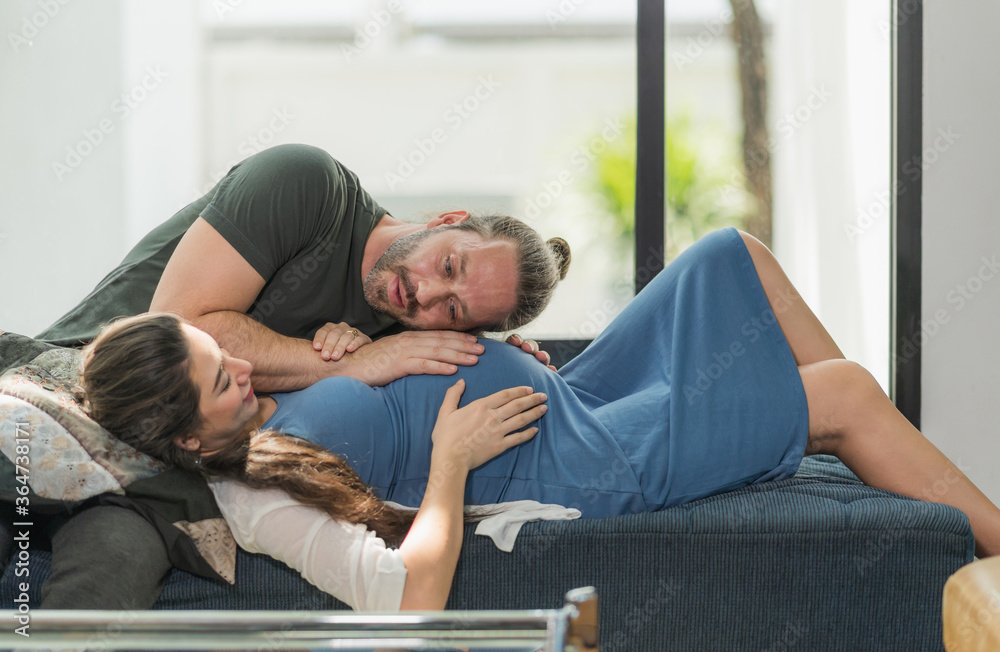 Beard man take care his pregnant wife lay down on sofa and listen to the baby.