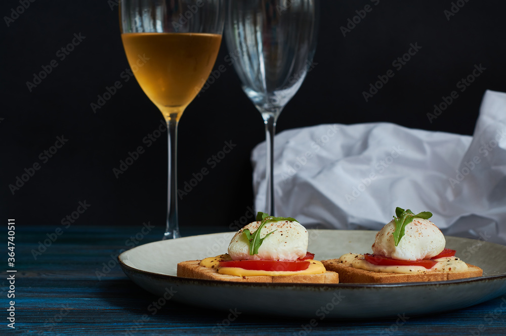 Large gray ceramic plate with toast and poached eggs, greens, tomatoes, sauce. Tasty breakfast. Glasses of wine. Large gray ceramic plate with toast and poached eggs, greens, tomatoes, sauce. Tasty breakfast. Glasses of wine.