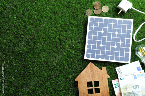 Flat lay composition with solar panel, house model and money on green grass. Space for text