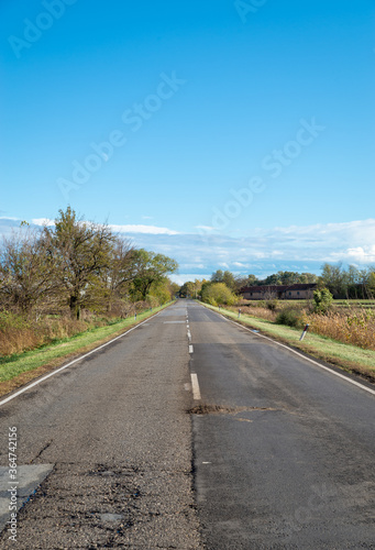 Cloudy country repaired highway/road in bad condition and blue cloudy sky. Green and yellow/orange fields, sunny landscape.