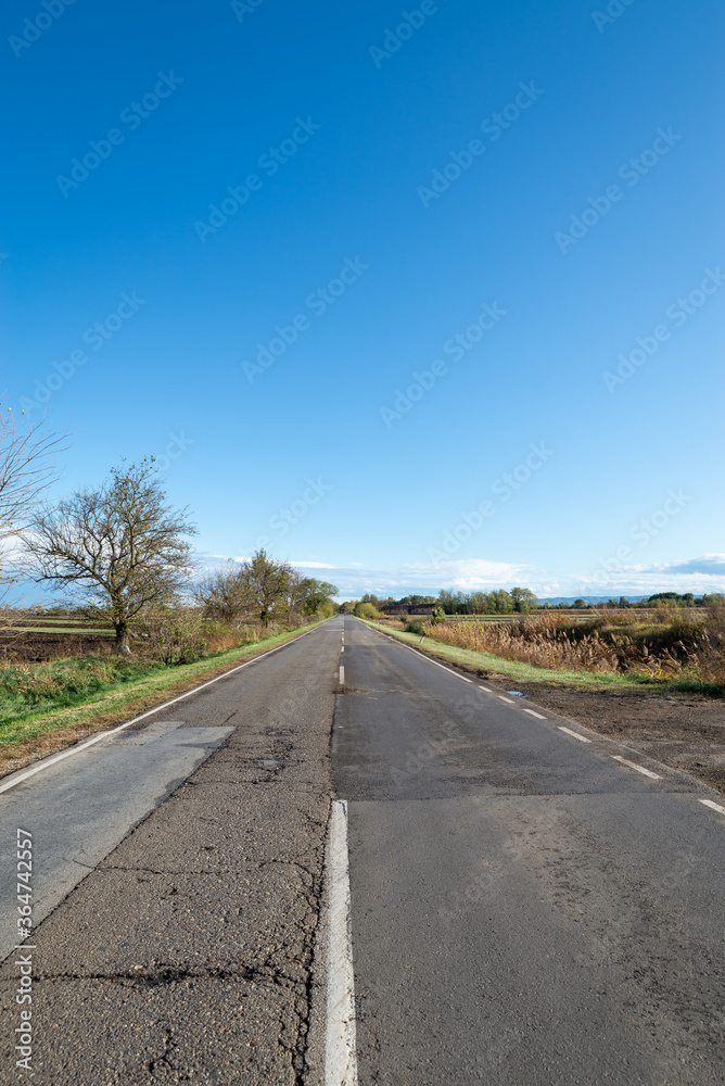 Cloudy country repaired highway/road in bad condition and blue cloudy sky. Green and yellow/orange fields, sunny landscape.