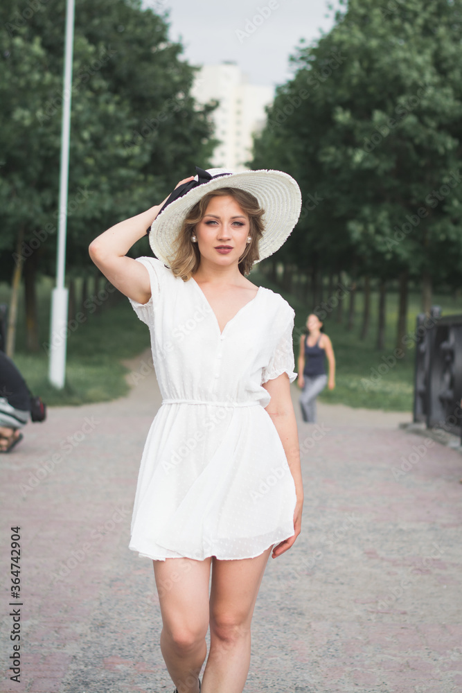 Stylish beautiful young girl in a white summer hat and a big white hat poses for a photographer on a footpath