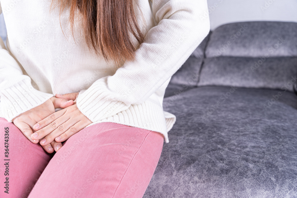Woman hands holding her crotch suffering from painful stomachache while sitting on sofa at home. Healthcare medical or daily life concept.