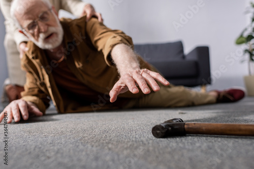 selective focus of fallen senior man trying to get walking stick while wife helping him