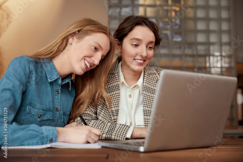 Students. Girls Studying With Laptop. Beautiful Young Women Having Lesson Online. Smiling Blonde And Brunette Looking At Screen And Writing Something.