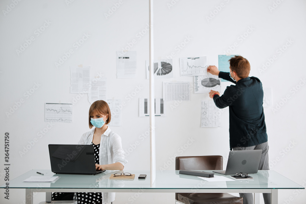Pretty girl and guy in medical masks working at their computers in the office. Coronavirus Covid-19. Social distancing.